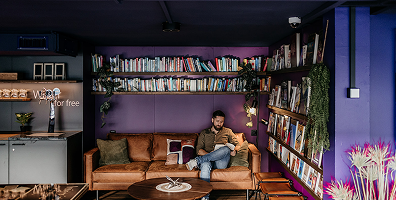 Capsule Hotel lounge library with purple walls; guest reading on leather sofa among bookshelves, plants, and warm lighting.