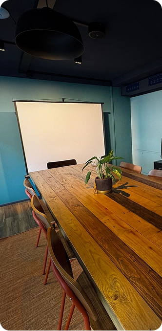 Capsule Hotel meeting room with long wooden table, potted plant centerpiece, chairs, and projection screen.