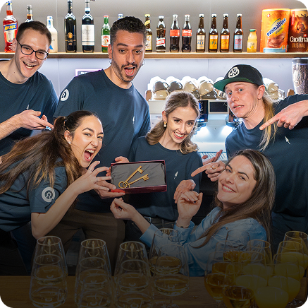 Capsule Hotel team celebrating behind bar, smiling and pointing at ceremonial keys amid glasses and bottle display