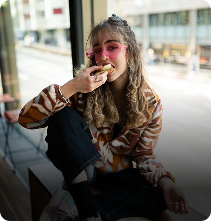Capsule Hotel guest with curly hair and pink glasses eats a pastry by the window in the lounge.