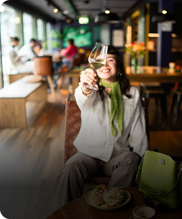 Smiling guest toasting with wine in the lively lounge at Capsule Hotel, with snacks, green bag, and warm decor.