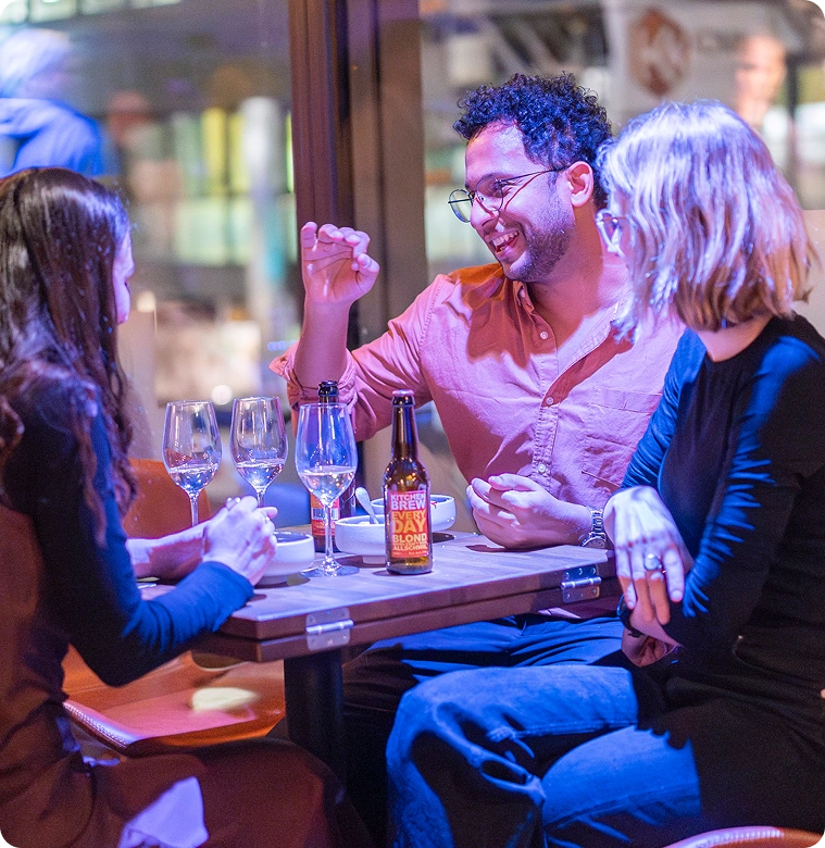 Capsule Hotel guests socializing over drinks in a modern lounge, with wine glasses and a beer bottle on the table.