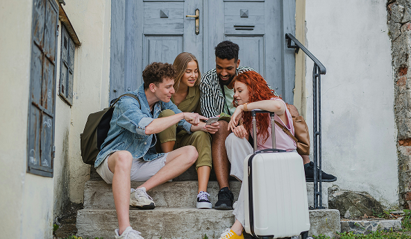 Four young travelers on stone steps checking a phone, with suitcase and backpacks, Capsule Hotel