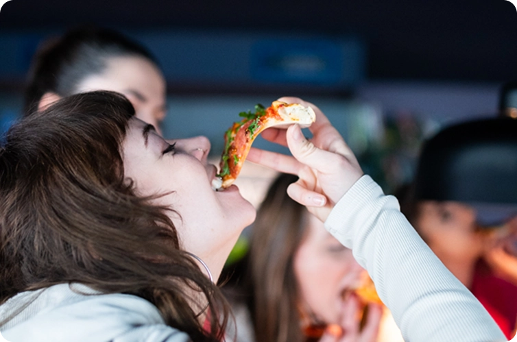 Capsule Hotel guest enjoys a slice of pizza in a lively social lounge, casual hangout vibe