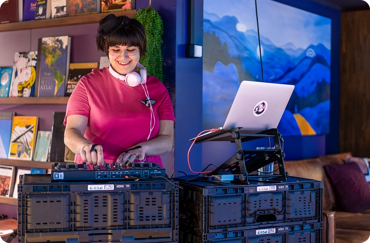 Capsule Hotel DJ in pink top mixing tracks on decks in a cozy lounge with bookshelves and blue artwork