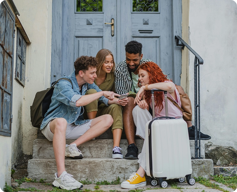 Capsule Hotel travelers: four friends on steps with suitcase, sharing a phone and laughing outside a city entrance.