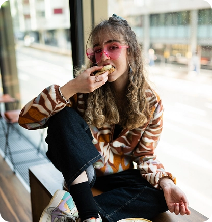 Capsule Hotel guest with curly hair and pink sunglasses eats a pastry by the window in a bright lounge.