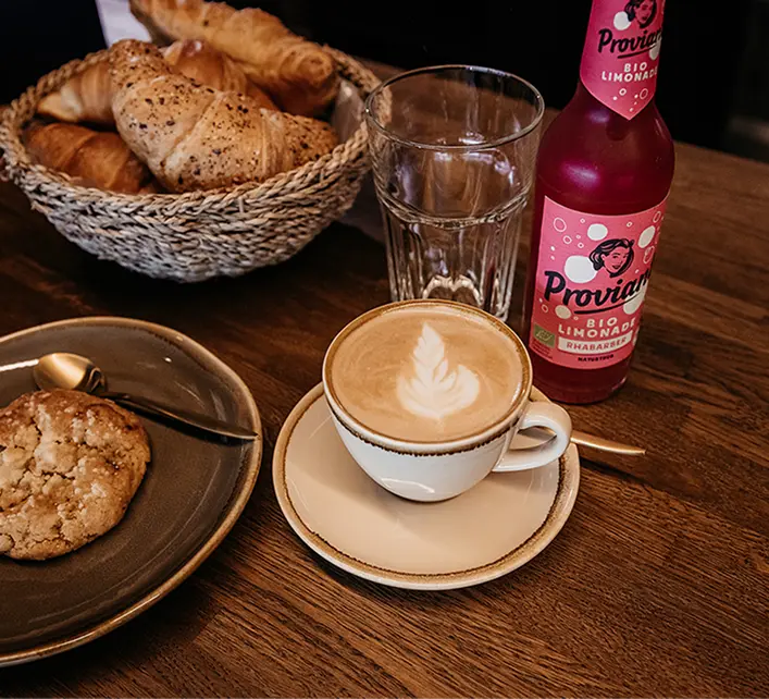 Capsule Hotel breakfast: latte with latte art, croissant basket, cookie on plate, glass, and pink Proviant lemonade.