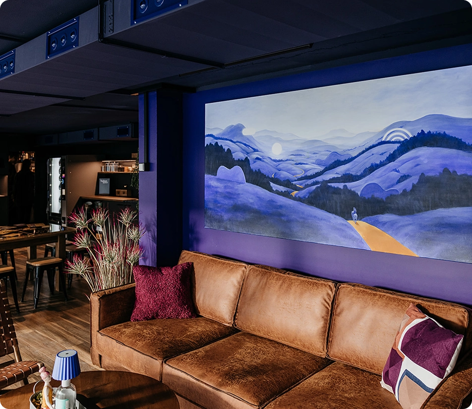 Capsule Hotel lounge with brown leather sofa, purple walls, and large blue landscape painting, wood tables and stools.
