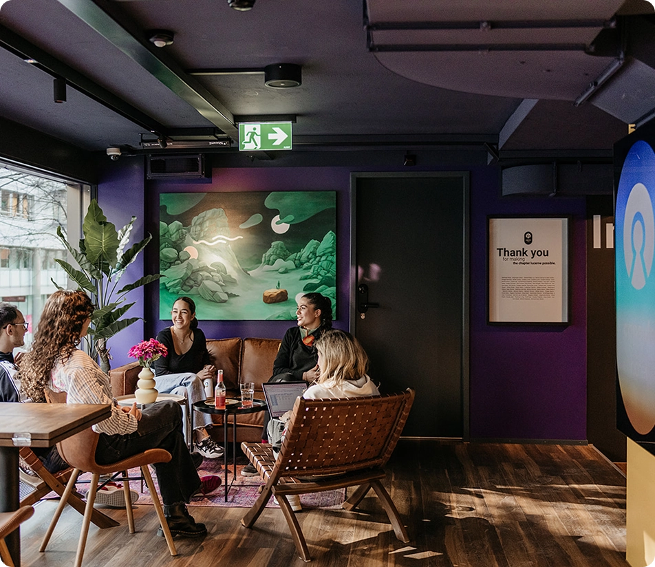 Capsule Hotel lounge with guests chatting on leather sofa, plants and art, cozy seating and warm lighting.
