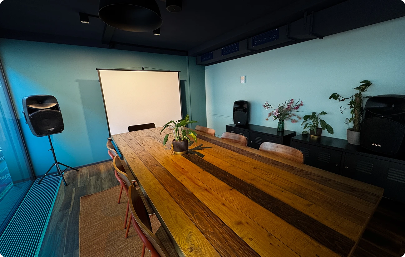 Capsule Hotel meeting room with long wooden table, chairs, projector screen, speakers, and potted plants.