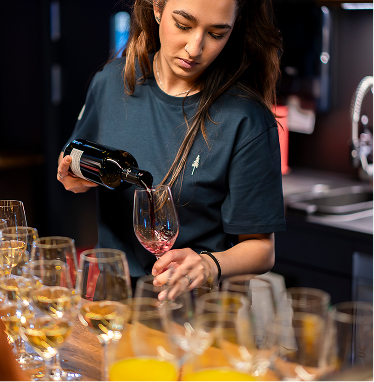 Capsule Hotel staff member pouring red wine at the bar, rows of glasses and juices in a modern lounge.