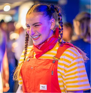 Capsule Hotel smiling person with braided hair in red overalls and striped tee under neon lights at a lively event