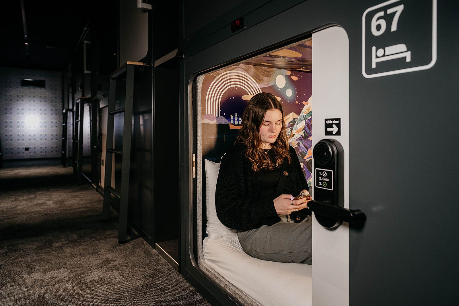 Inside capsule room with a woman using a phone at Capsule Hotel Lucerne Old Town, featuring a space-themed wall design.