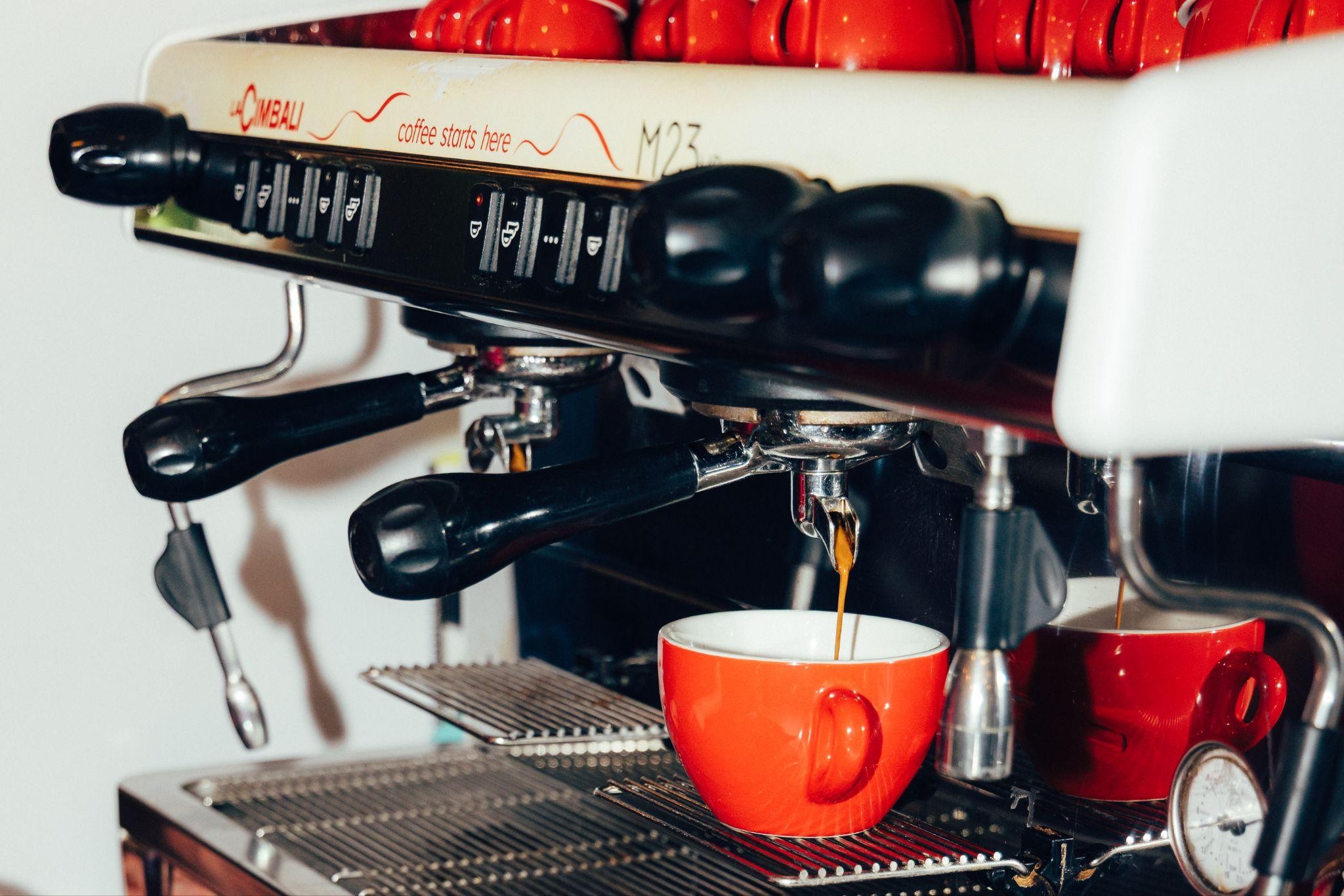 Capsule Hotel espresso machine pouring a shot into a red cup, with red mugs stacked on top in the café.