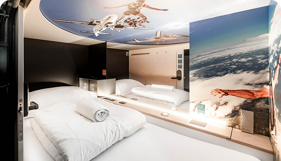 Capsule Hotel modern pod with twin beds, soft LED lighting, and sky-themed mural with skydivers and clouds.