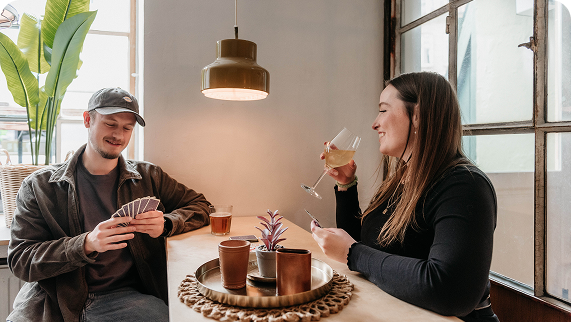 Two guests playing cards in cozy lounge with drinks, pendant lamp, and window at Capsule Hotel