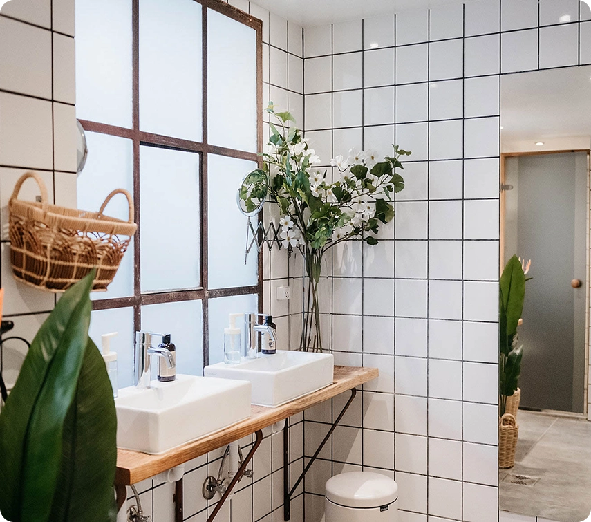 Capsule Hotel tiled bathroom with double sinks, frosted window, greenery, and wicker accents
