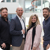 Capsule Hotel team of four standing in a bright modern atrium, smiling in smart-casual attire.