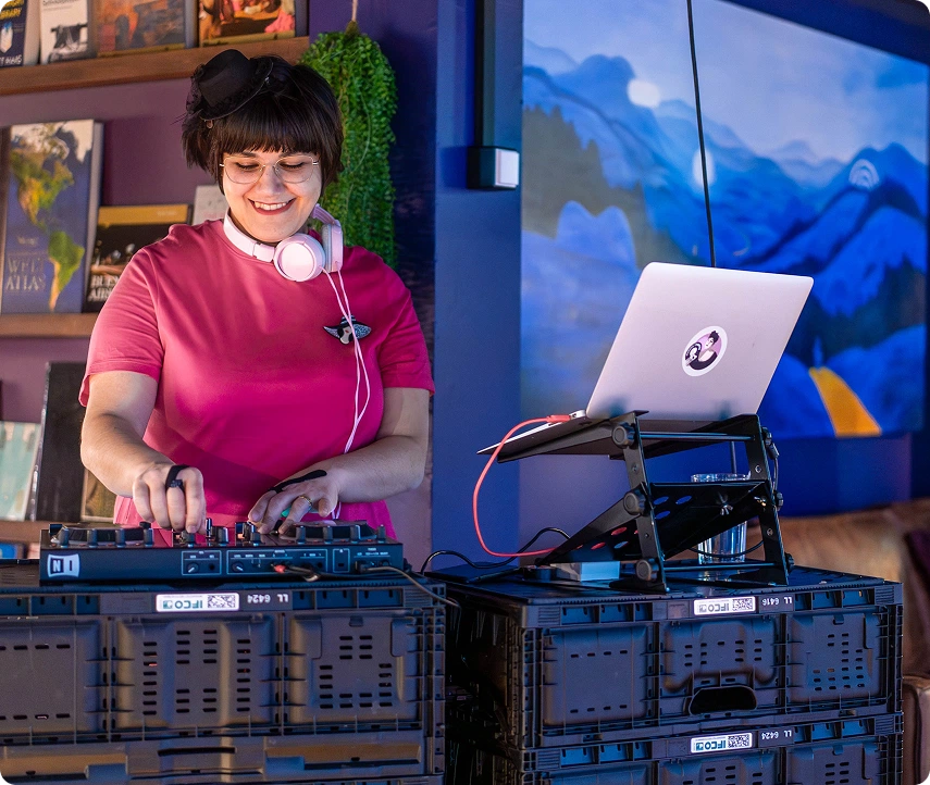 DJ in pink shirt mixing tracks in Capsule Hotel lounge, headphones on, laptop and decks set on crates, blue mural behind