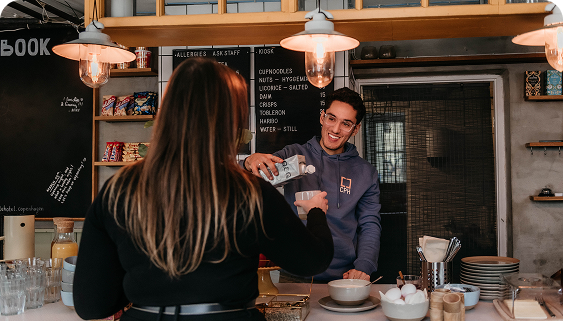 Capsule Hotel staff member pours milk for guest at cozy breakfast bar with chalkboard menu and warm pendant lights.