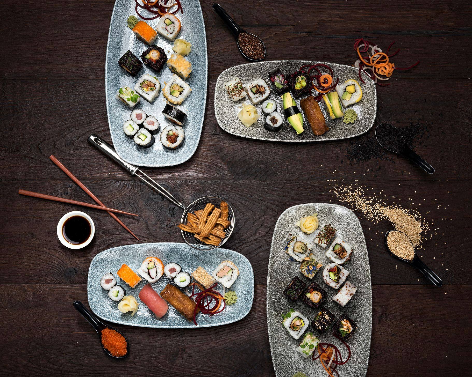 Sushi-Platten im Capsule Hotel auf einem dunklen Holztisch mit Essstäbchen, Sojasauce und verstreuten Sesamsamen.
