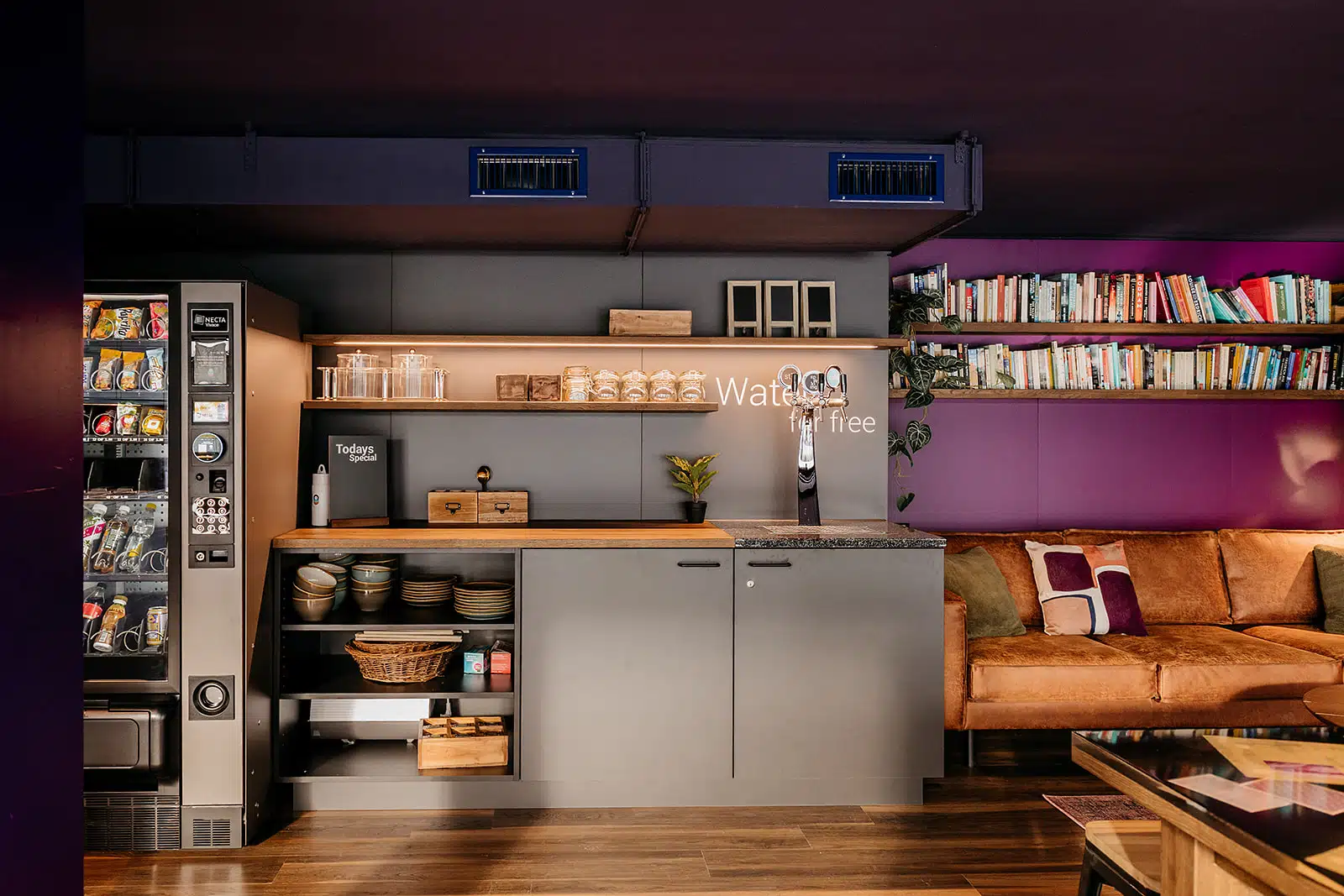 Cozy common area at Capsule Hotel with a vending machine, bookshelves, and a plush sofa for guests.