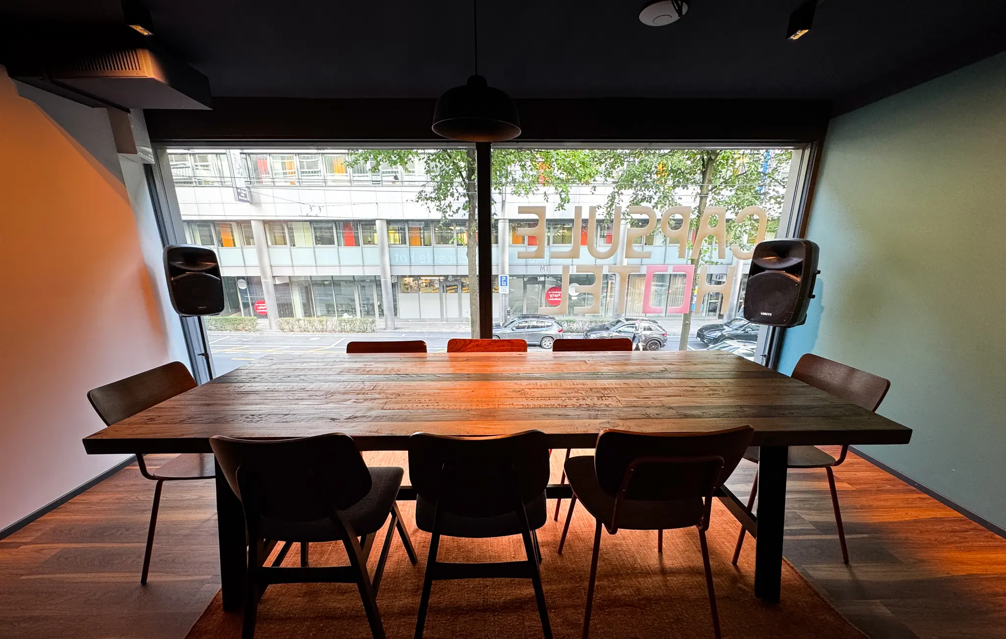 Capsule Hotel meeting room with long rustic table, eight chairs, speakers, and street view through large window.