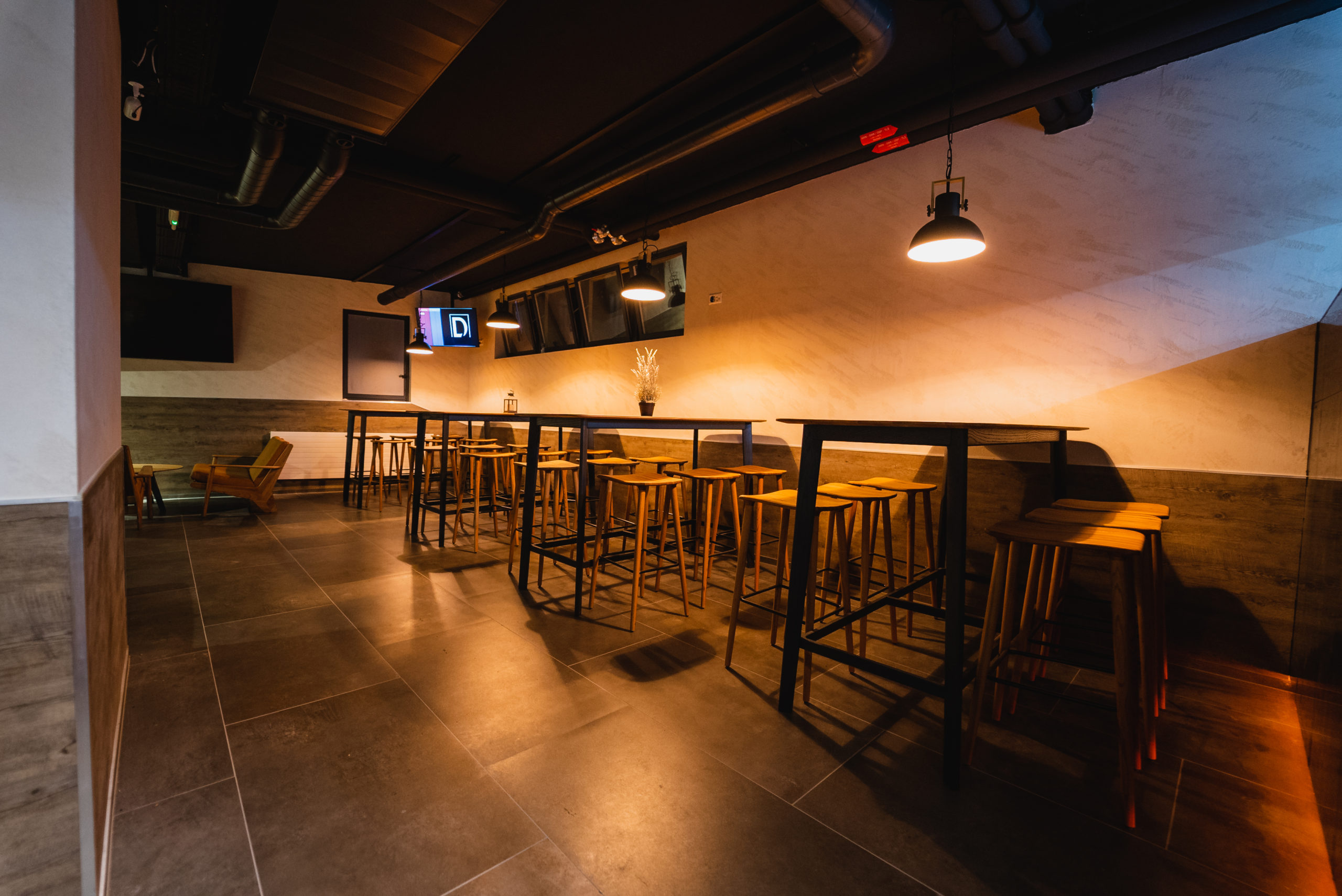 Moderne Lounge im Capsule Hotel mit hohen Tischen, Holzschemeln, Hängelampen und einer industriellen Decke in warmem Licht.