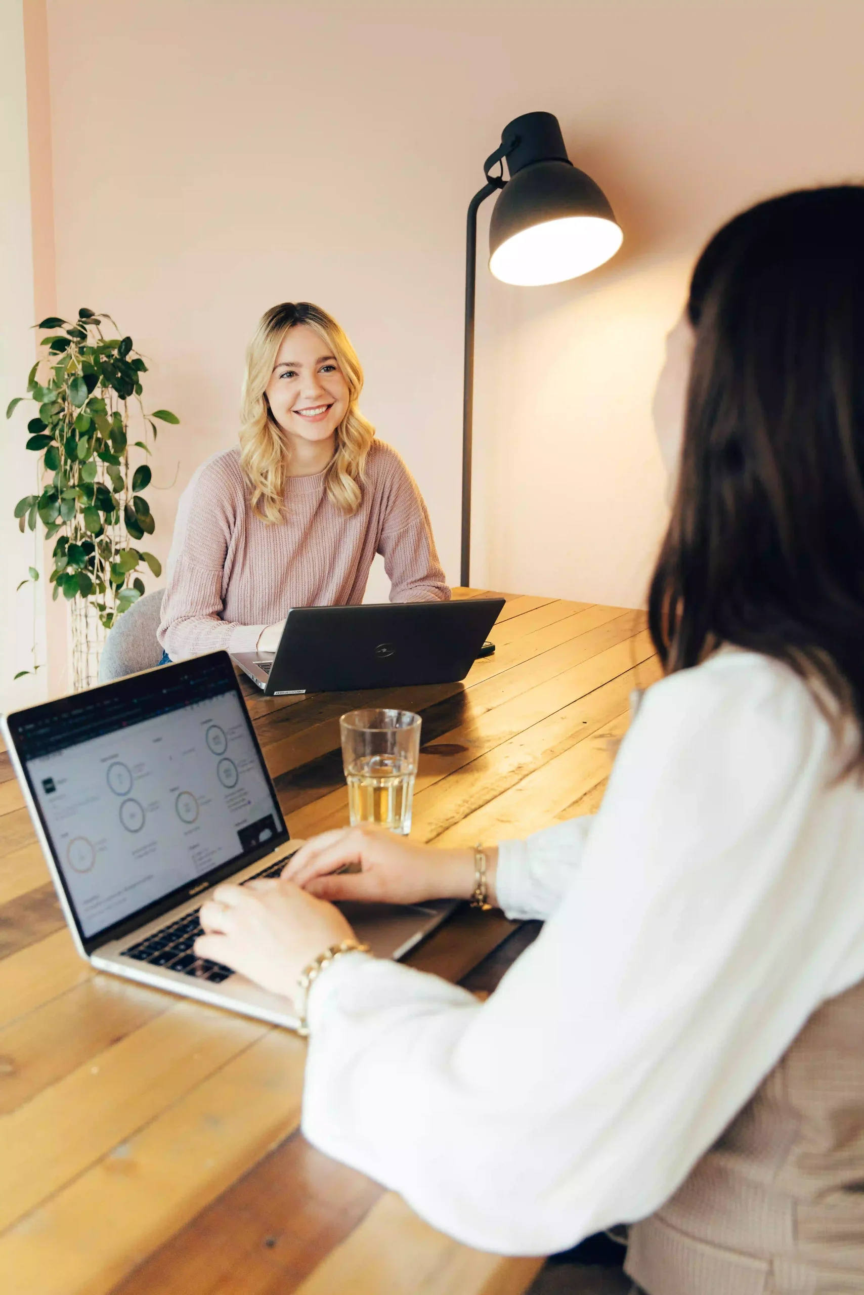 Team-Meeting im Capsule Hotel: zwei Frauen mit Laptops an einem Holztisch, eine warme Lampe und eine Pflanze in einem modernen Arbeitsbereich.
