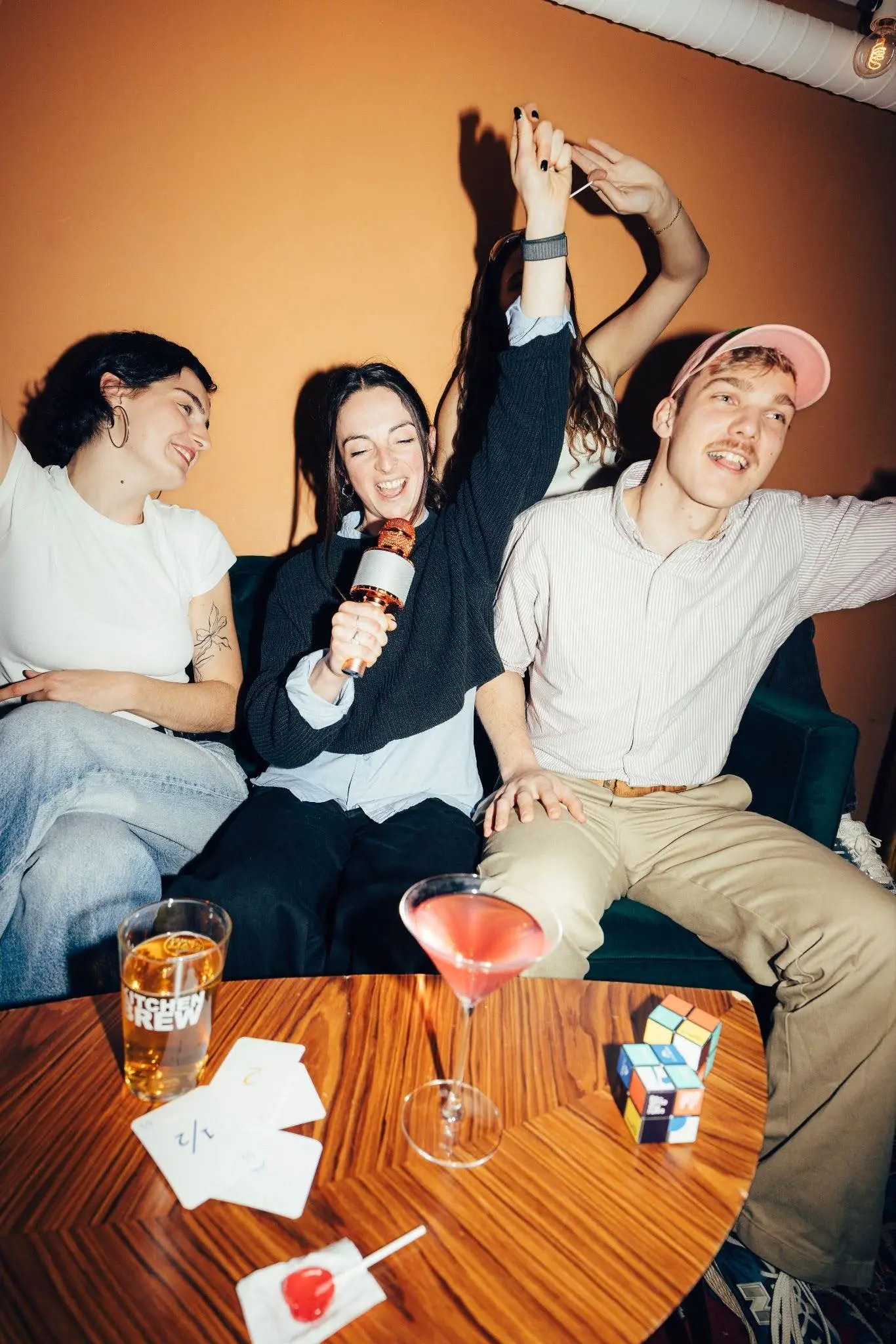 Kapselhotel-Gäste singen Karaoke auf einem Sofa, lachen, mit Cocktails, Karten und einem Rubik's Cube auf einem Holztisch.