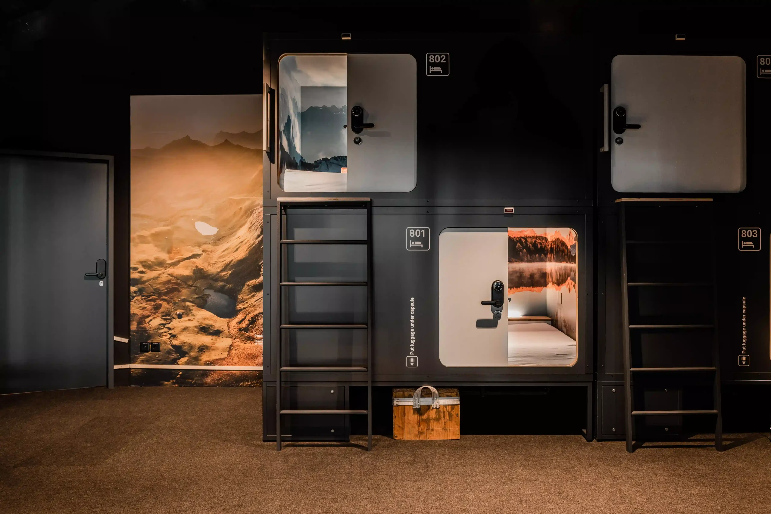 Capsule Hotel stacked sleeping pods 801–803 with ladders and cozy beds beside a warm mountain landscape mural.