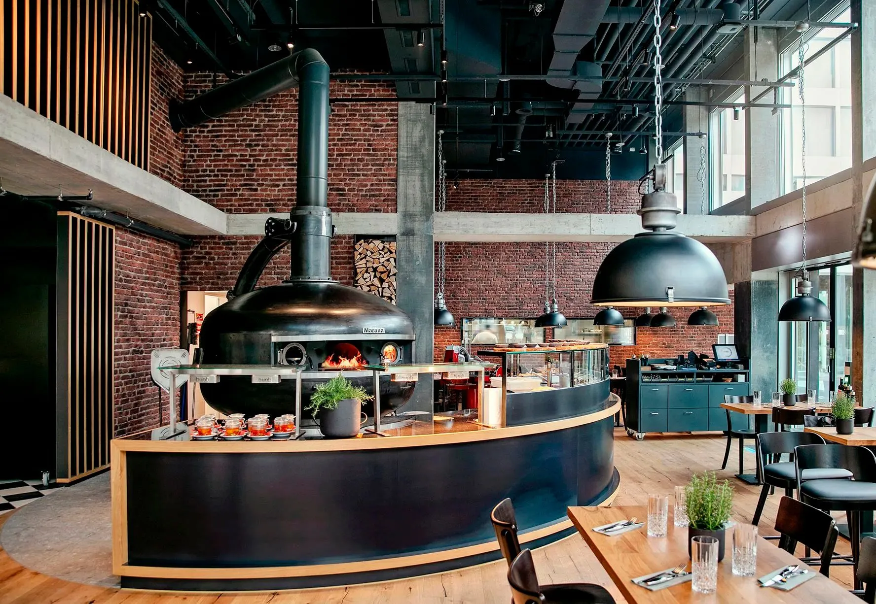 Industrial-Chic Restaurant im Capsule Hotel mit Holzofen, Ziegelwänden, schwarzen Hängeleuchten und Holztischen.