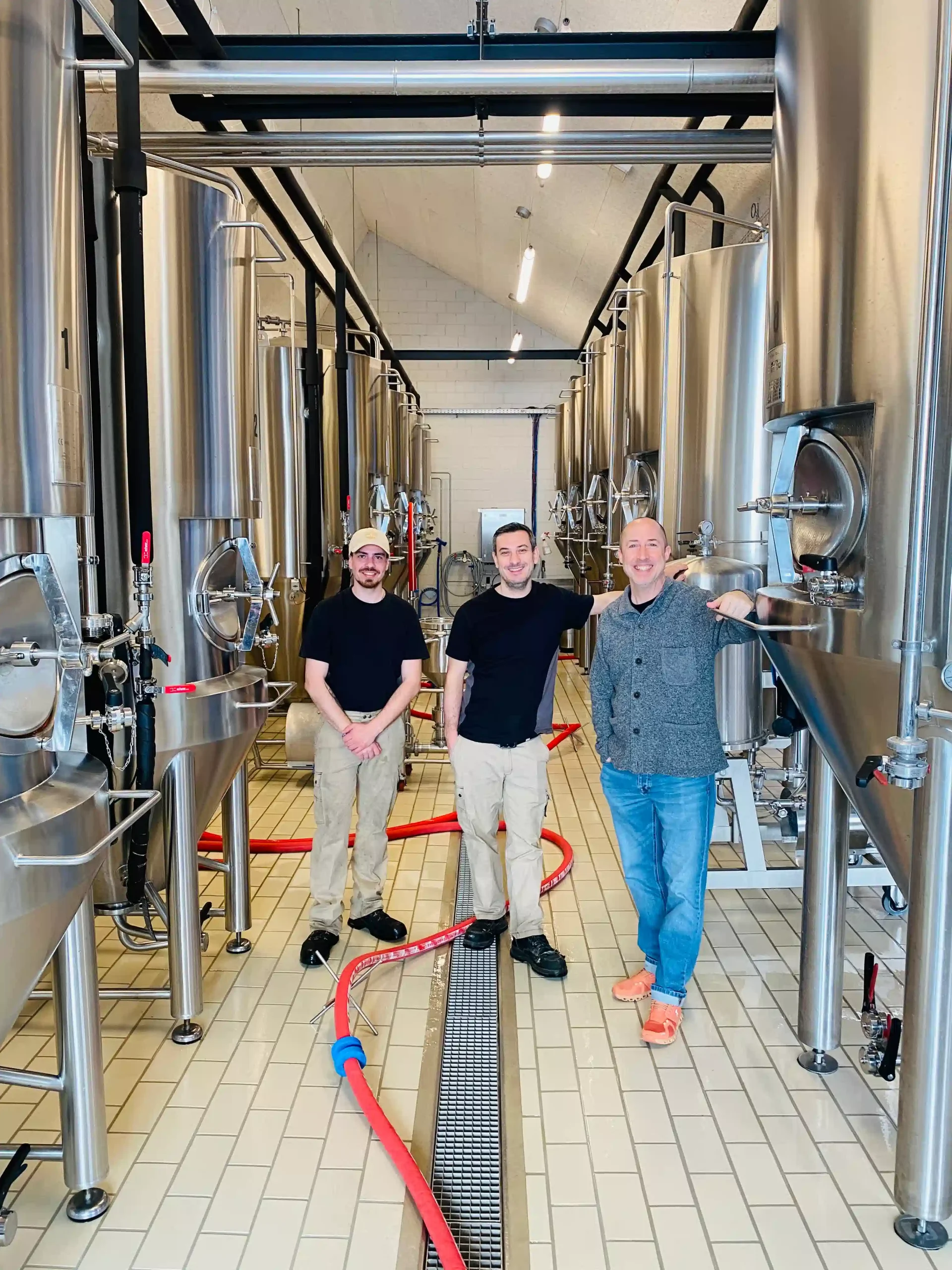 Three Capsule Hotel team members between stainless steel brewing tanks, red hose on tiled floor in bright brewery.