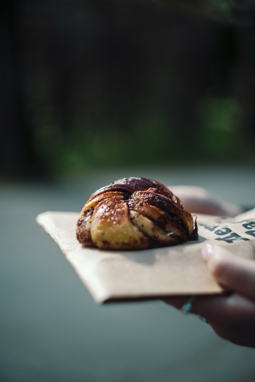 Eine Capsule Hotel Zimtschnecke auf Kraftpapier, draußen in der Hand gehalten, ein glänzendes geflochtenes Gebäck zum Frühstück.