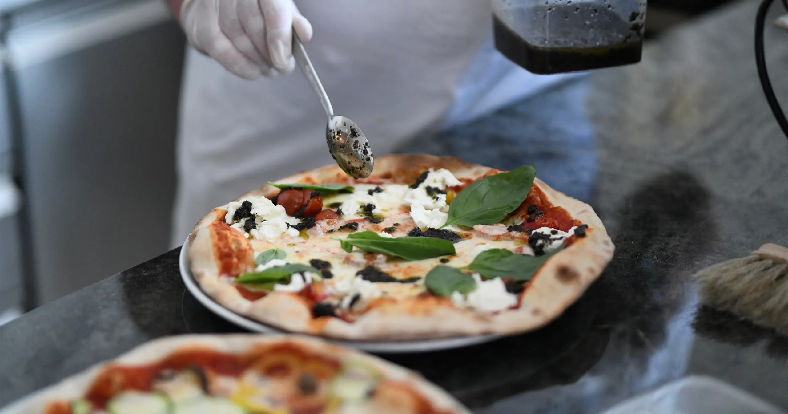Kapselhotel-Koch mit Handschuhen, der eine Steinofenpizza mit Spinat, Ricotta und Oliventapenade in der Küche fertigstellt