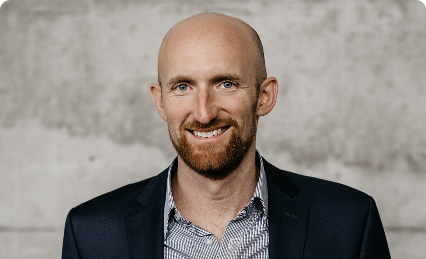 Capsule Hotel team portrait: smiling bald man with ginger beard in navy blazer against a concrete wall backdrop.