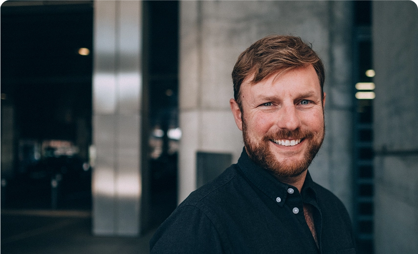 Capsule Hotel staff portrait: smiling bearded man in a dark shirt against a modern industrial backdrop