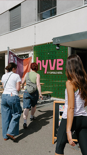 People walking toward Hyve by Capsule Hotel entrance, green sign and streetfront at 64