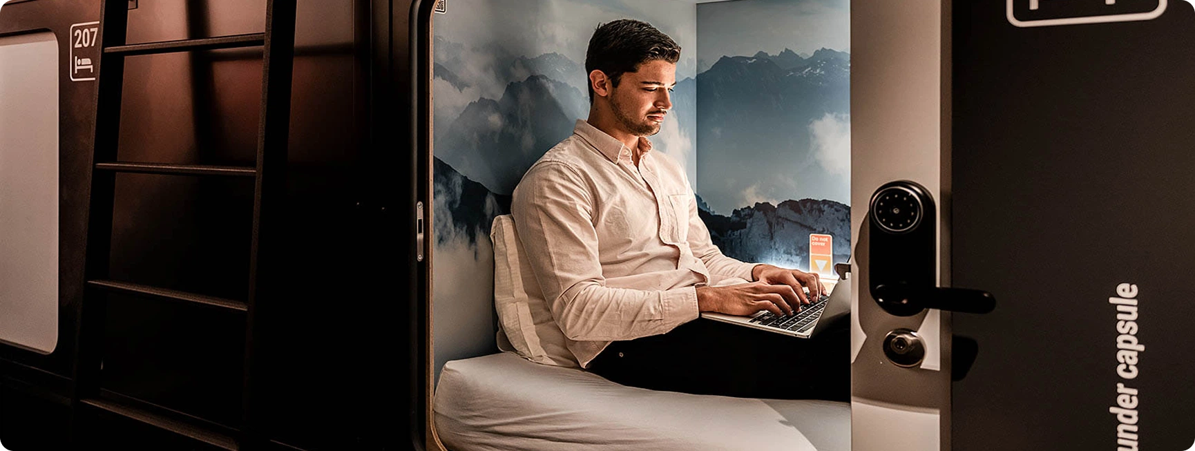 Capsule Hotel guest working on laptop in private capsule bed with mountain mural; pod door and ladder visible