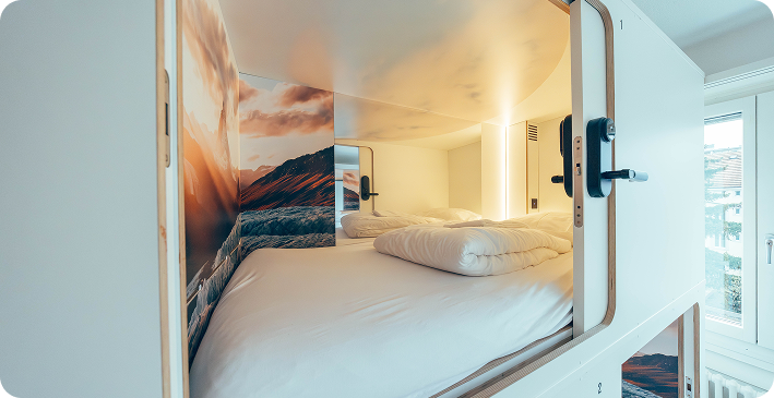 Capsule Hotel cozy pod with white bedding, reading light, and mountain mural, bright natural light from nearby window.