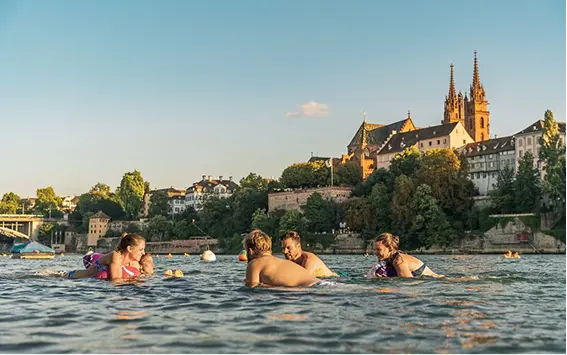 Families swimming in a scenic European river near historic architecture, promoting the adventure spirit of Capsule Hotel.