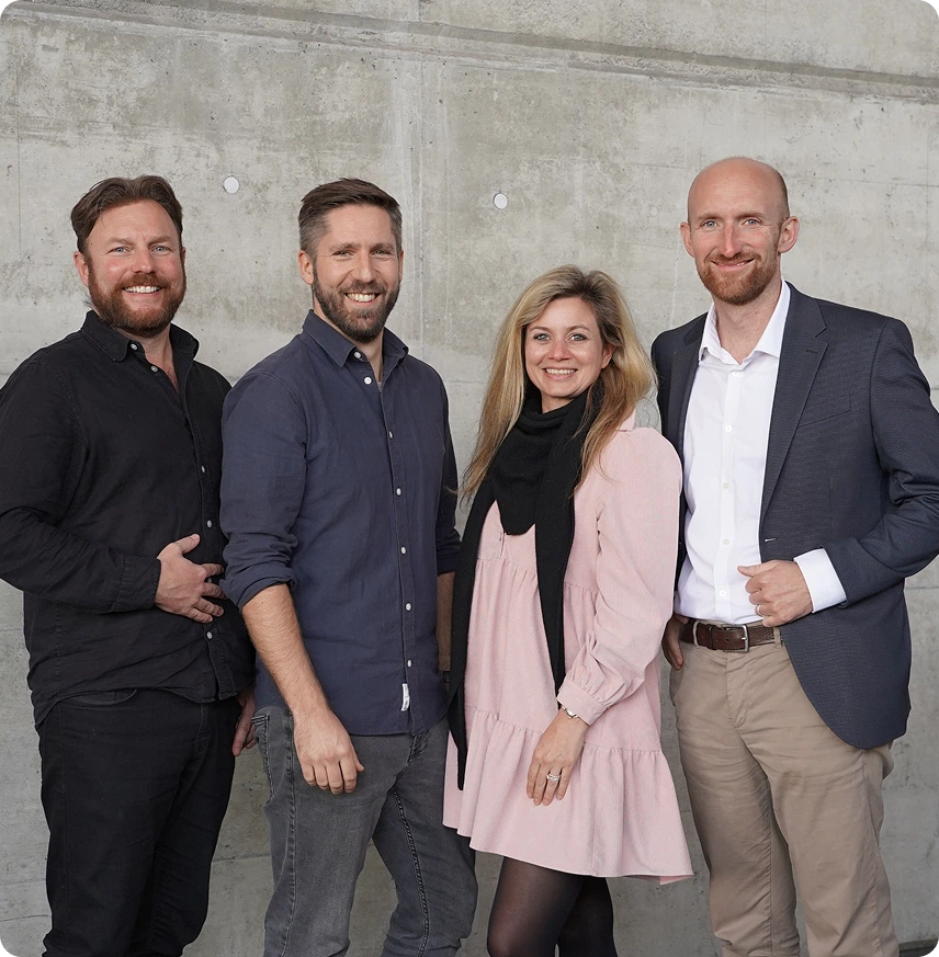 Capsule Hotel leadership team, four smiling colleagues by a concrete wall in smart-casual outfits