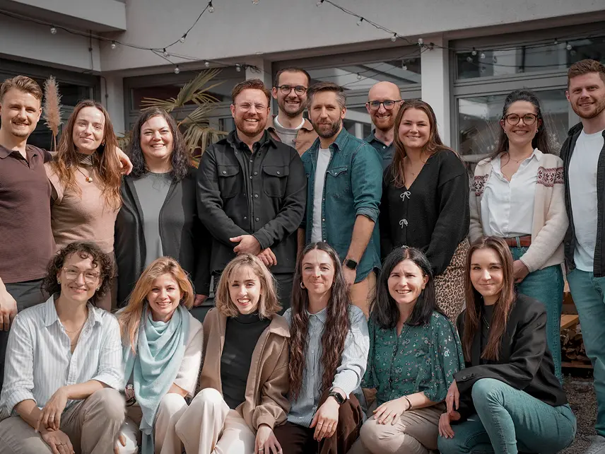 Capsule Hotel team group smiling in a sunny courtyard, casual attire, standing and seated under string lights