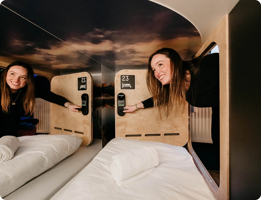 Two smiling women testing smart locks on adjacent sleeping pods at Capsule Hotel, beds with rolled towels.