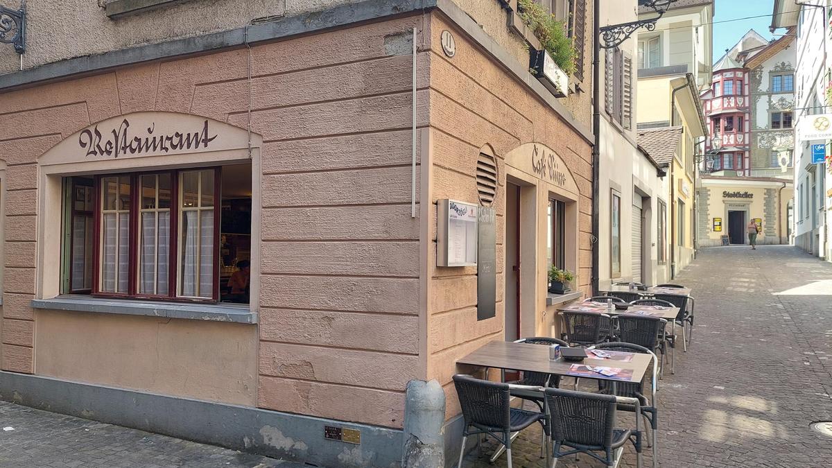 Europäische Altstadtstraße am Capsule Hotel mit gemütlicher Café-Terrasse, Kopfsteinpflaster und pastellfarbenen Fassaden im sanften Tageslicht.