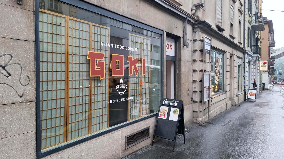 Straßenszene im Viertel des Capsule Hotels mit dem Goki Asian Food Laden und nassem Gehweg an einem regnerischen Tag.