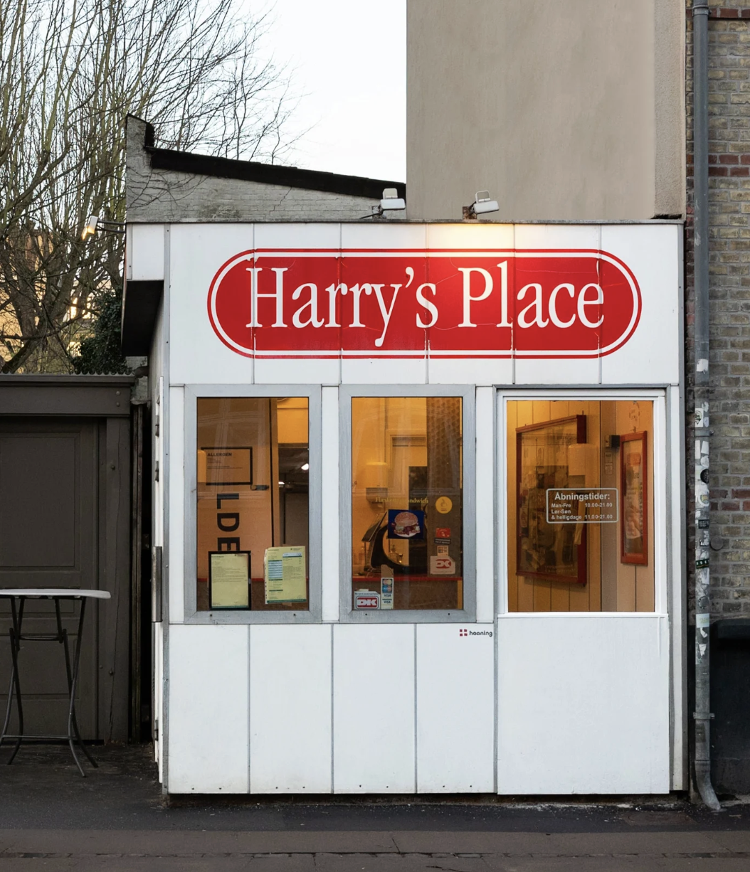 Städtische Straßenszene am Capsule Hotel mit Harry’s Place Kiosk, auffälligem roten Schild und warm beleuchteten Fenstern.