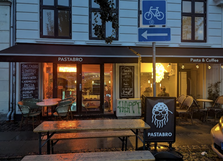 Städtische Szene am Capsule Hotel mit der Außenansicht des Pastabro Cafés in der Dämmerung, nassen Bänken, Neonlicht und gemütlichen Sitzgelegenheiten im Freien.