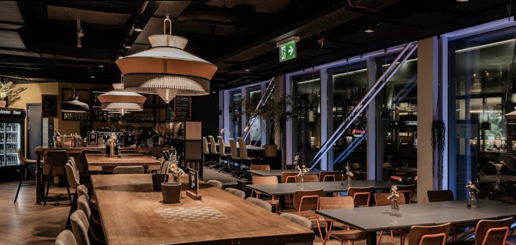 Lounge und Bar im Capsule Hotel mit warmen Hängeleuchten, langen Holztischen und Fenstern mit Stadtblick bei Nacht.